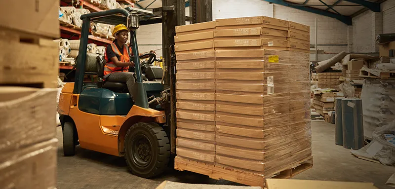 Young African female forklift operator moving boxes on a pallet around the floor of a storehouse.